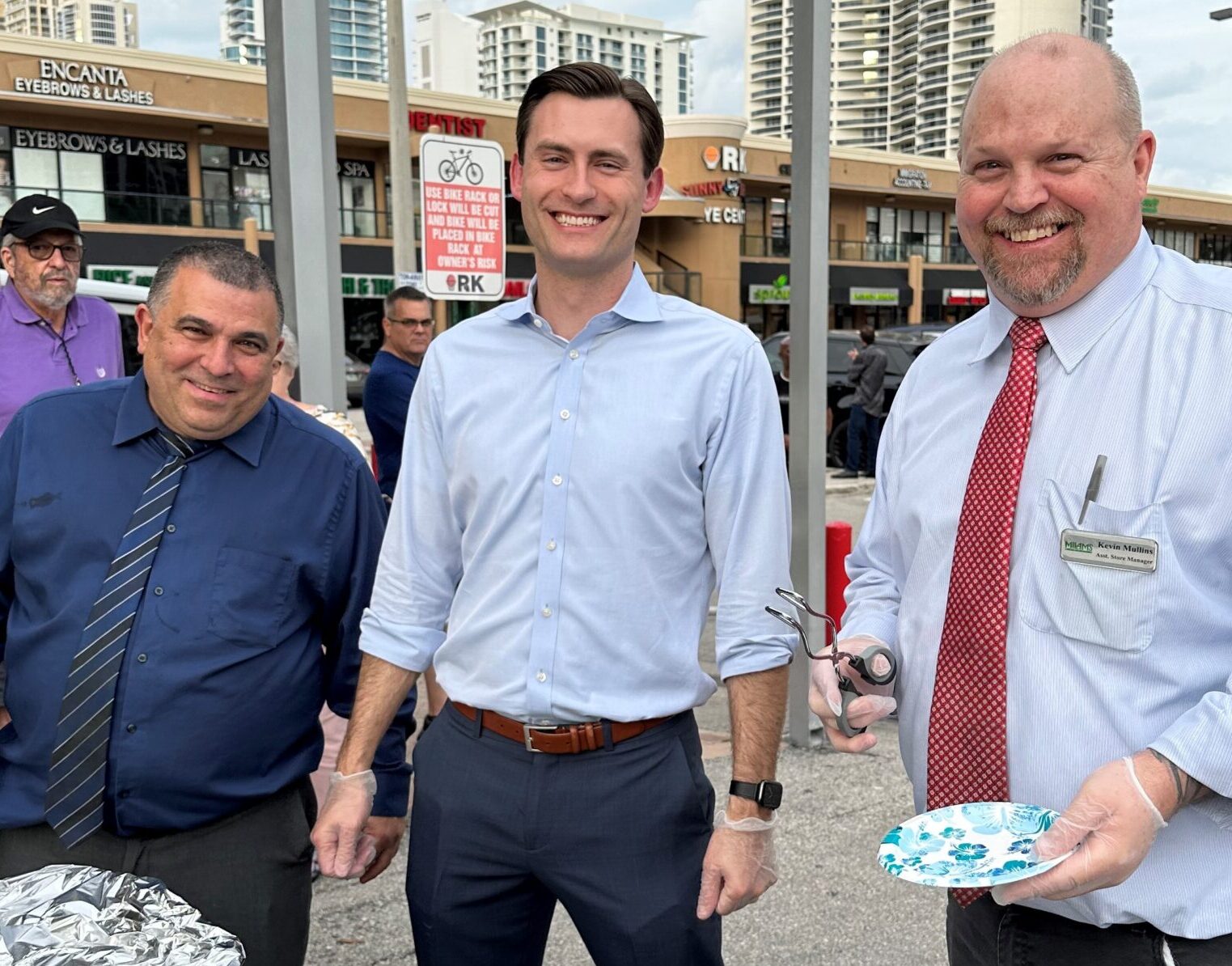 Three men stand outdoors, smiling at the camera. Dressed in business attire and wearing gloves, they hold plates and serving utensils, highlighting their involvement in a Community Donations event. Buildings and shops are visible in the background.