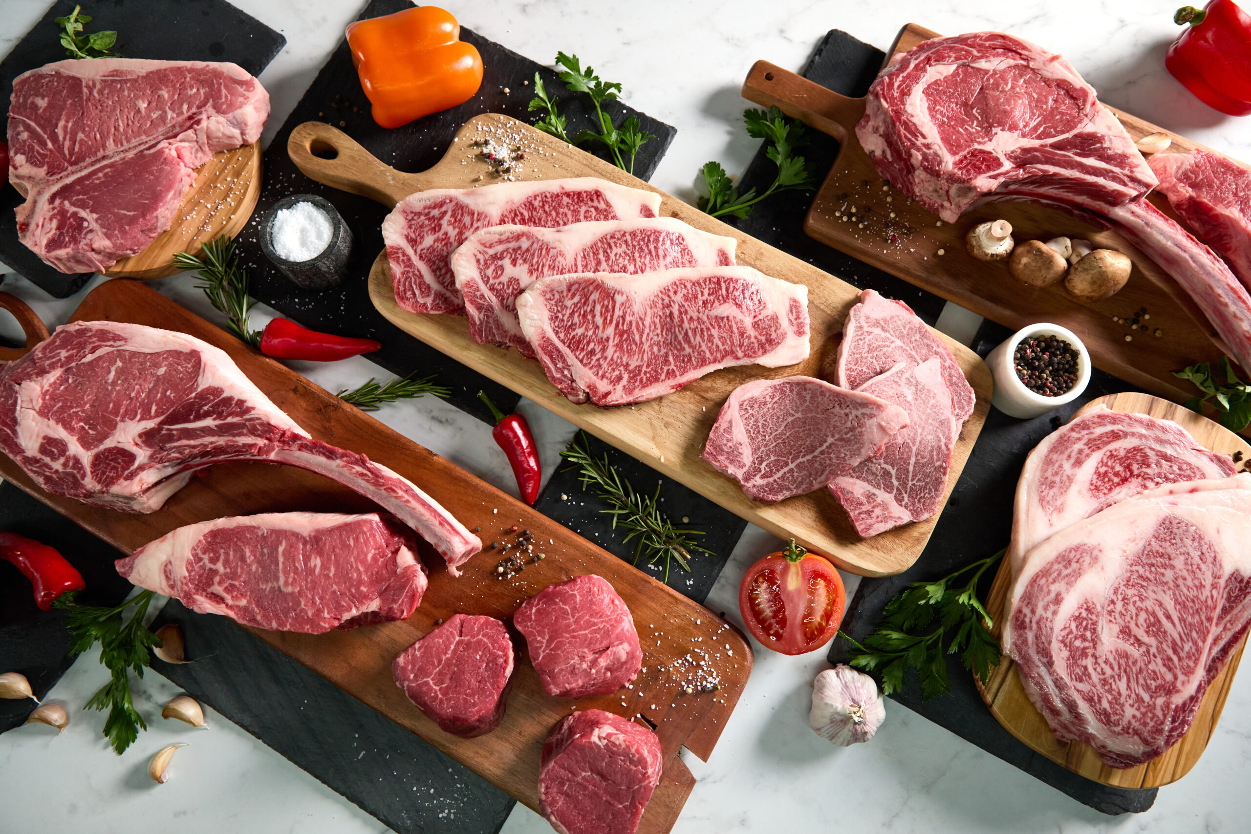 A premium meat selection featuring fresh cuts of marbled steak is artfully arranged on wooden boards with herbs, spices, tomatoes, garlic, mushrooms, and peppers atop a white marble surface—just like at a high-end butcher counter.