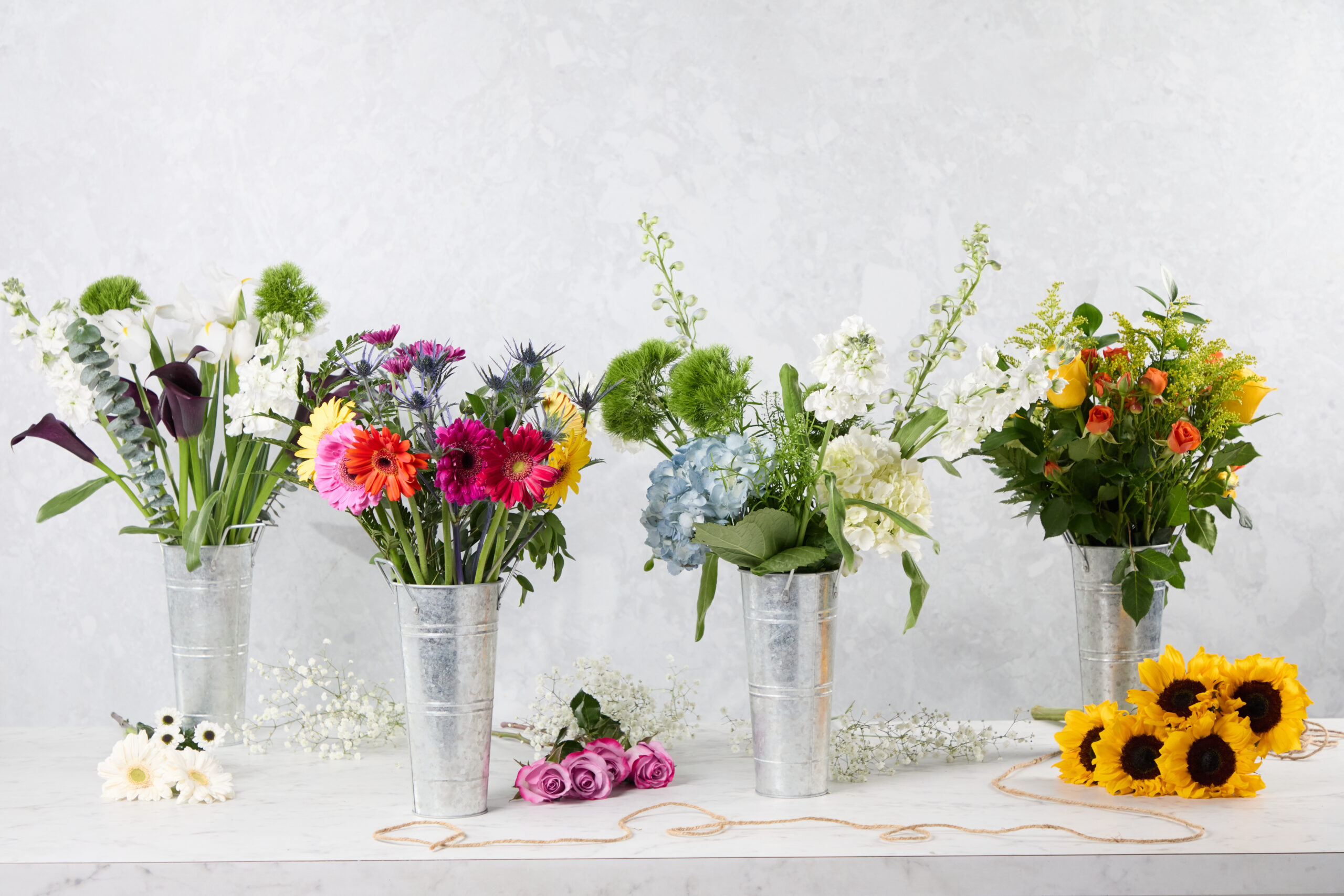 Five metal vases with assorted vibrant floral arrangements—sunflowers, roses, gerbera daisies, and hydrangeas—are arranged on a white marble surface with loose favorite florals and greenery scattered around.