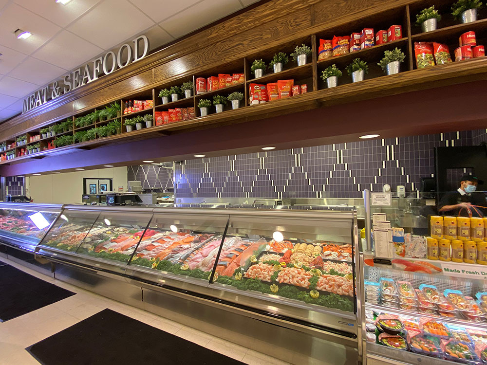 A grocery store meat and seafood counter displays various fresh meats and Fresh Seafood. Above the counter are shelves with potted plants and food products, and a sign that reads MEAT & SEAFOOD.