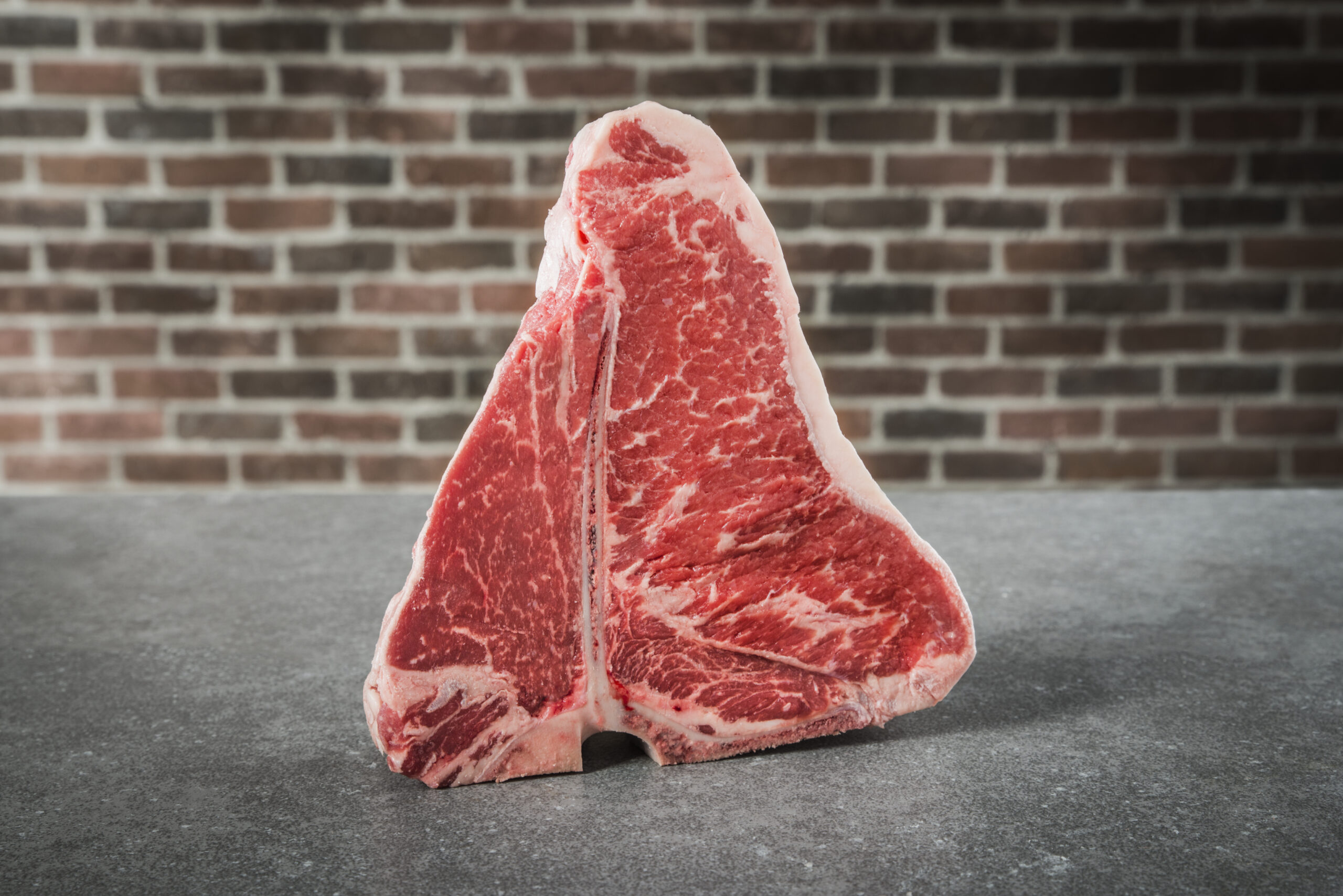 A raw T-bone steak stands upright on a gray countertop, with a red and gray brick wall backdrop. Featuring visible marbling and its classic T-shaped bone, it’s a striking example for any beef cuts explained or featured in a beef guide.