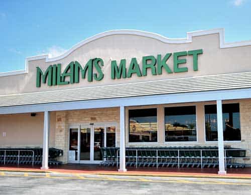 The entrance of Milams Market, featuring green lettering on the building facade, glass double doors, and rows of shopping carts lined up outside under a sunny sky.