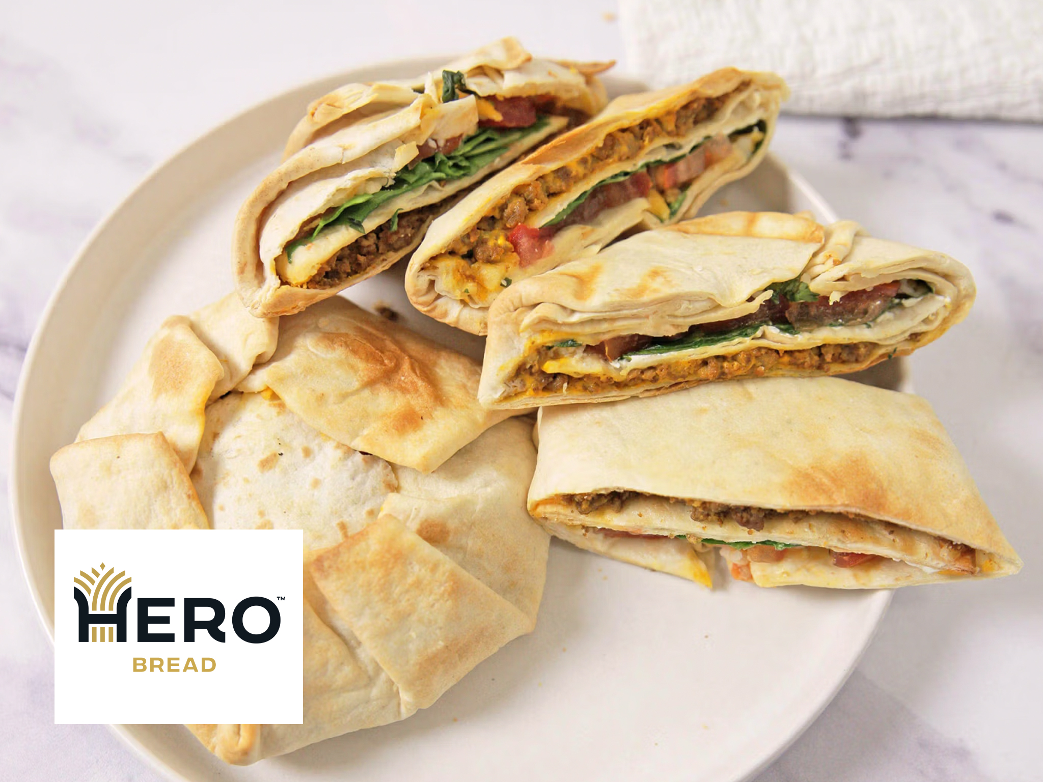 Three folded Crunchwraps filled with eggs, vegetables, and greens are served on a white plate—one whole wrap and two halves. A white napkin and the HERO Bread logo appear in the foreground. Perfect for an easy Air Fryer breakfast!.