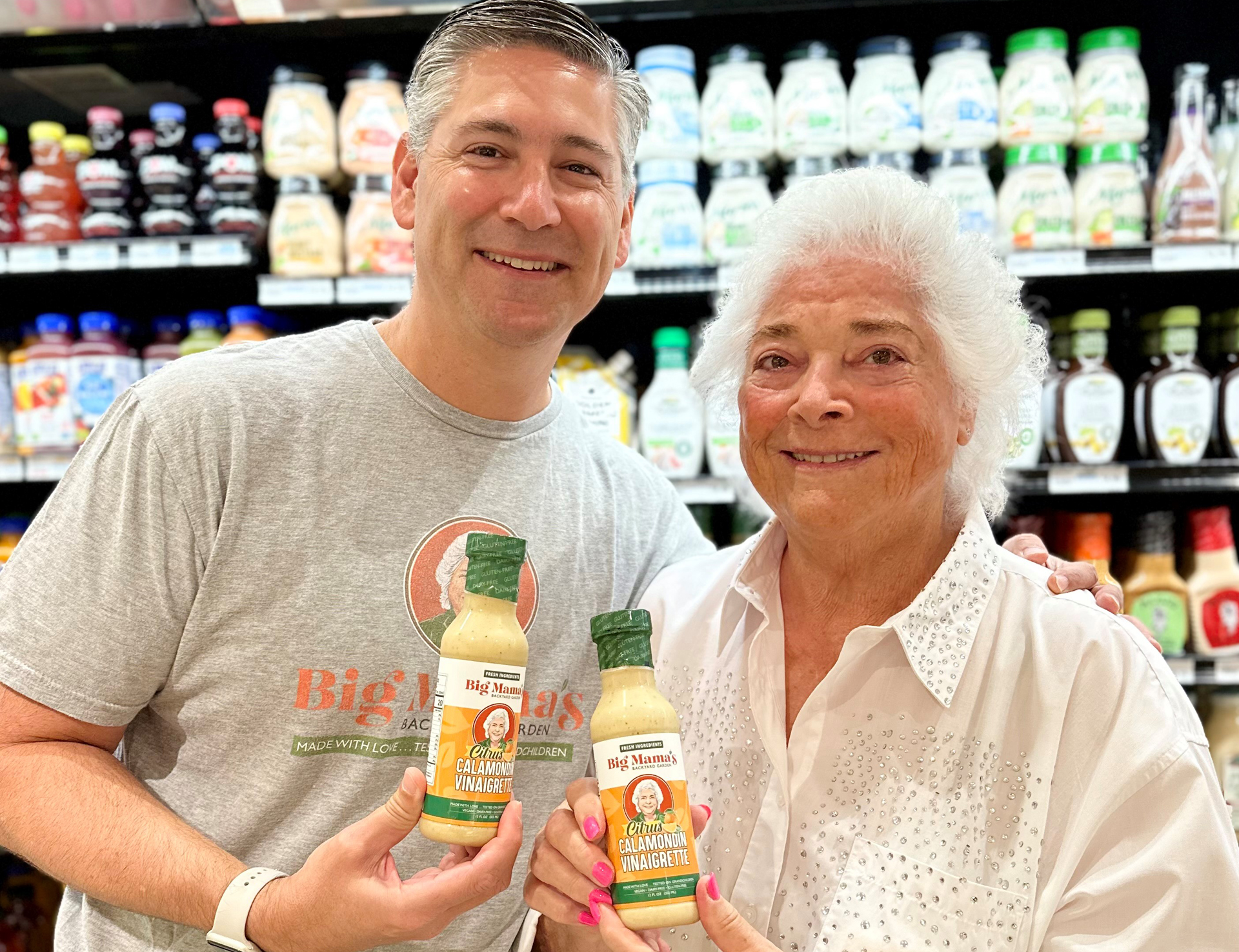 A smiling man and woman stand in a grocery store aisle holding bottles of Big Mama's salad dressing, with rows of Calamondin vinaigrette and other dressings visible on the shelves behind them.