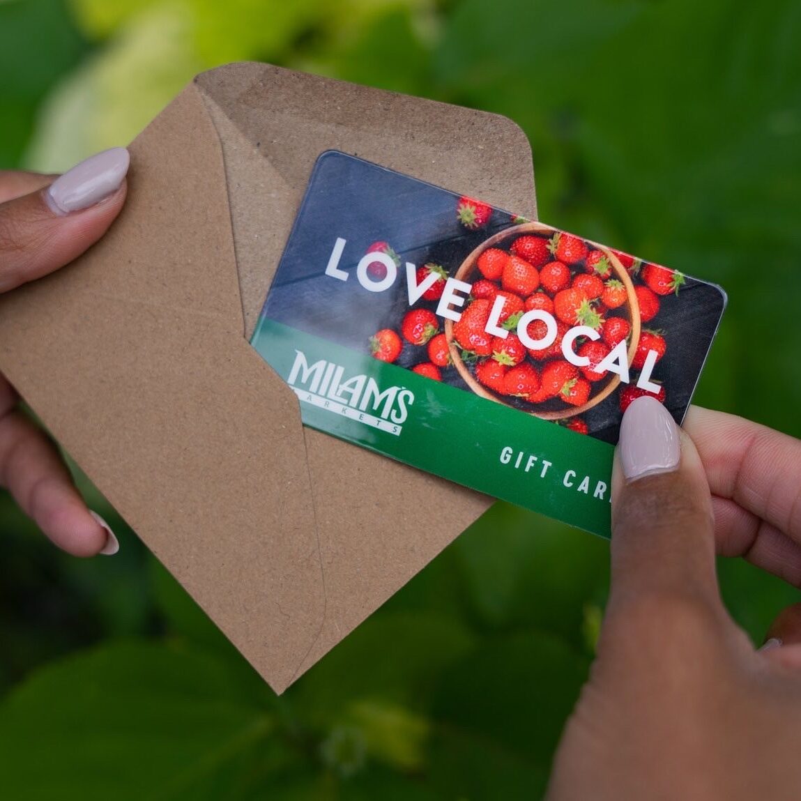 A person holds a brown envelope with a Love Local gift card partially pulled out. The card, featuring strawberries and "Milams Market Gift Card" text against green, highlights Order Online convenience. Green leaves are seen in the background.
