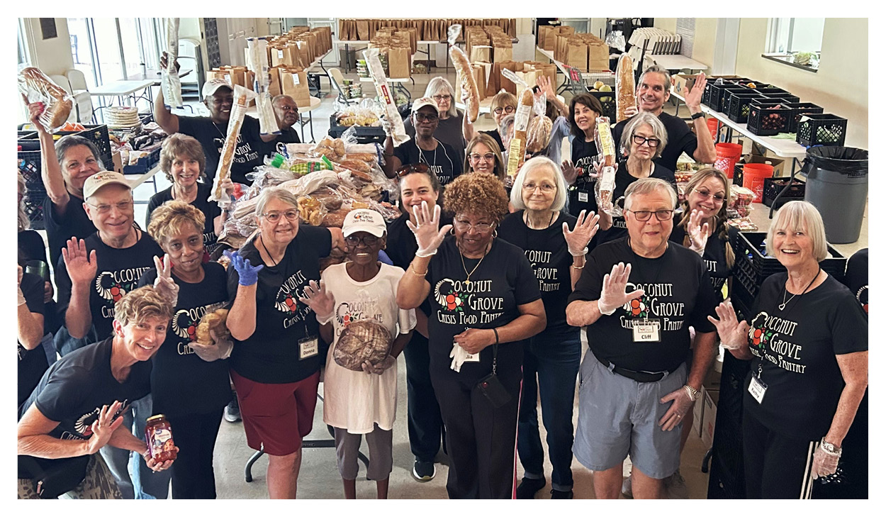A group of smiling volunteers wearing matching t-shirts stand together in a large room filled with tables of food, waving at the camera during a local food banks fundraising event.