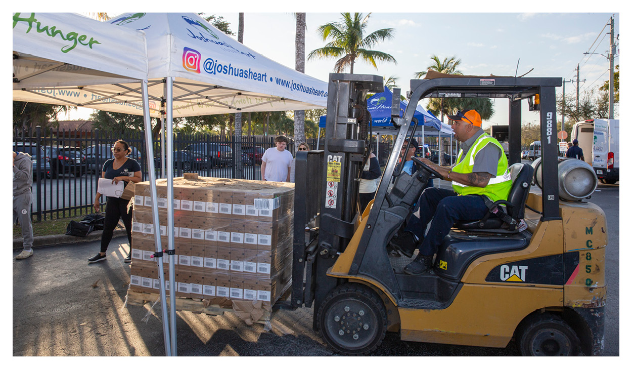 A man operates a forklift to move a pallet stacked with boxes under a white tent labeled “Hunger,” supporting local food banks. Several people stand nearby, with palm trees and a fence visible in the background.
