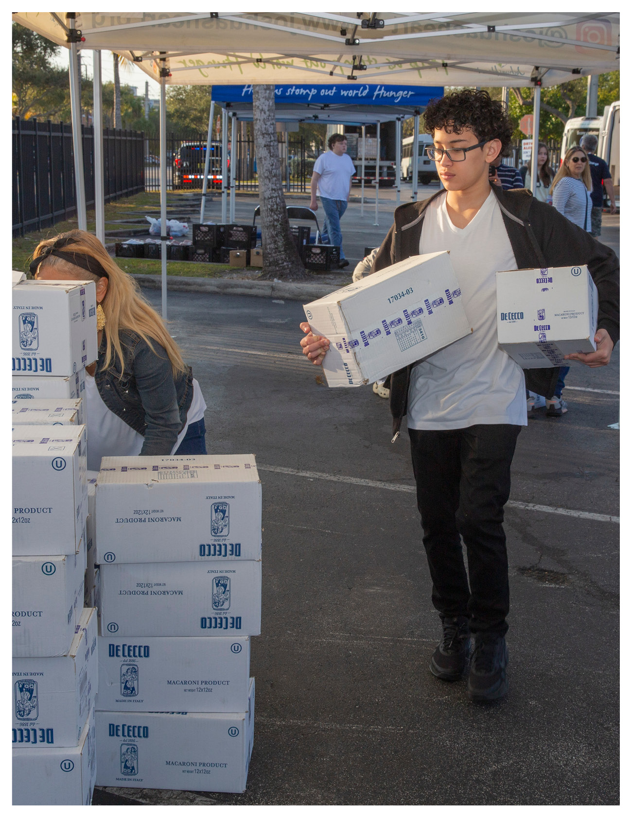 A young man carrying a box of De Cecco pasta walks past stacked boxes at a charity event, while a woman nearby bends over to pick up another box. A tent and vehicles are visible in the background, highlighting support for local food banks.