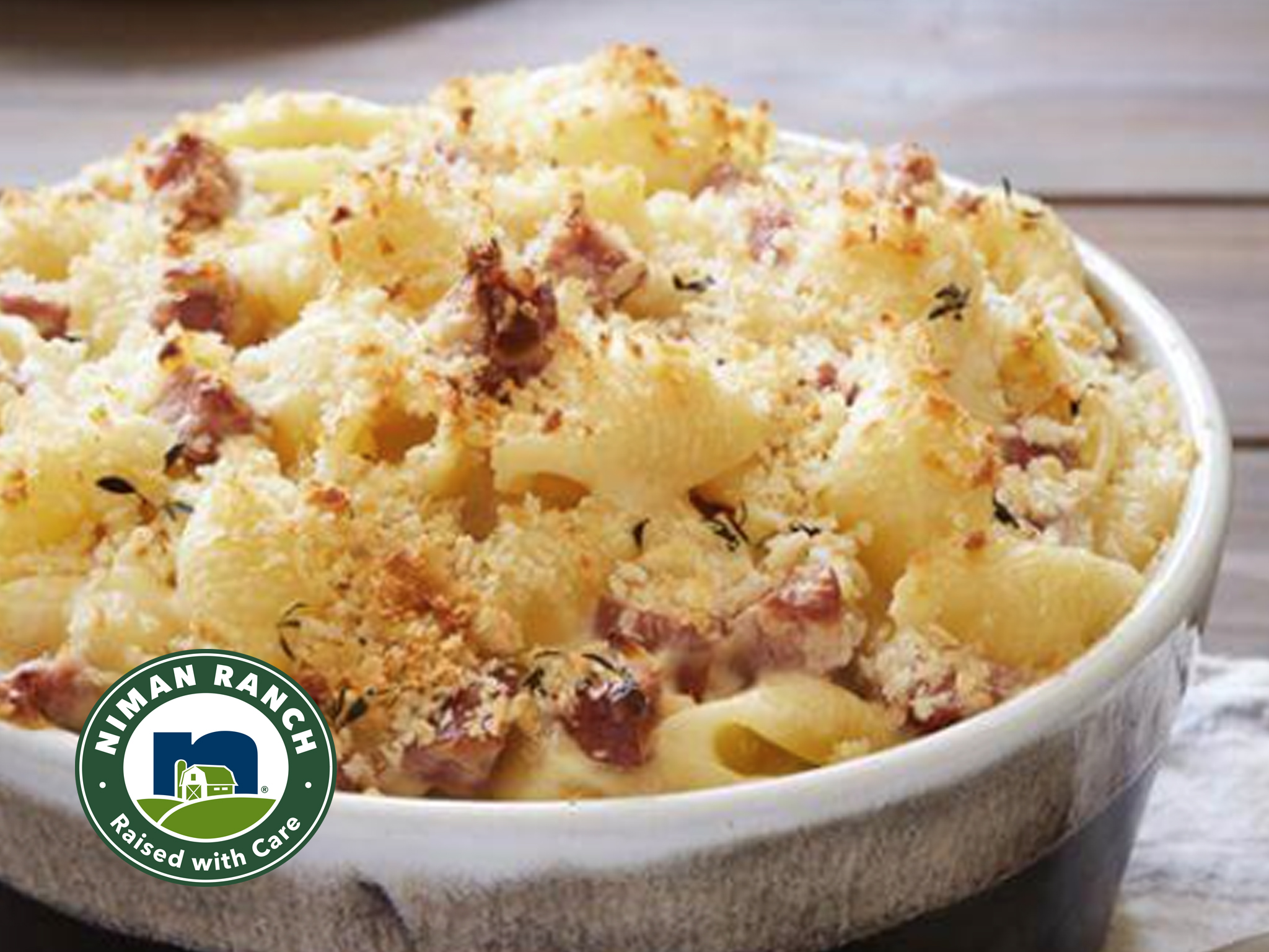 A Baked Macaroni & Cheese dish topped with breadcrumbs and bits of Apple Gouda Sausage is shown in a white casserole dish. The Niman Ranch logo appears in the bottom left corner.