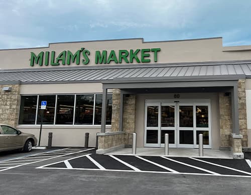Exterior view of Milam’s Market storefront with large green sign, glass doors, accessible parking spaces, and a beige car parked to the left under a partly cloudy sky.