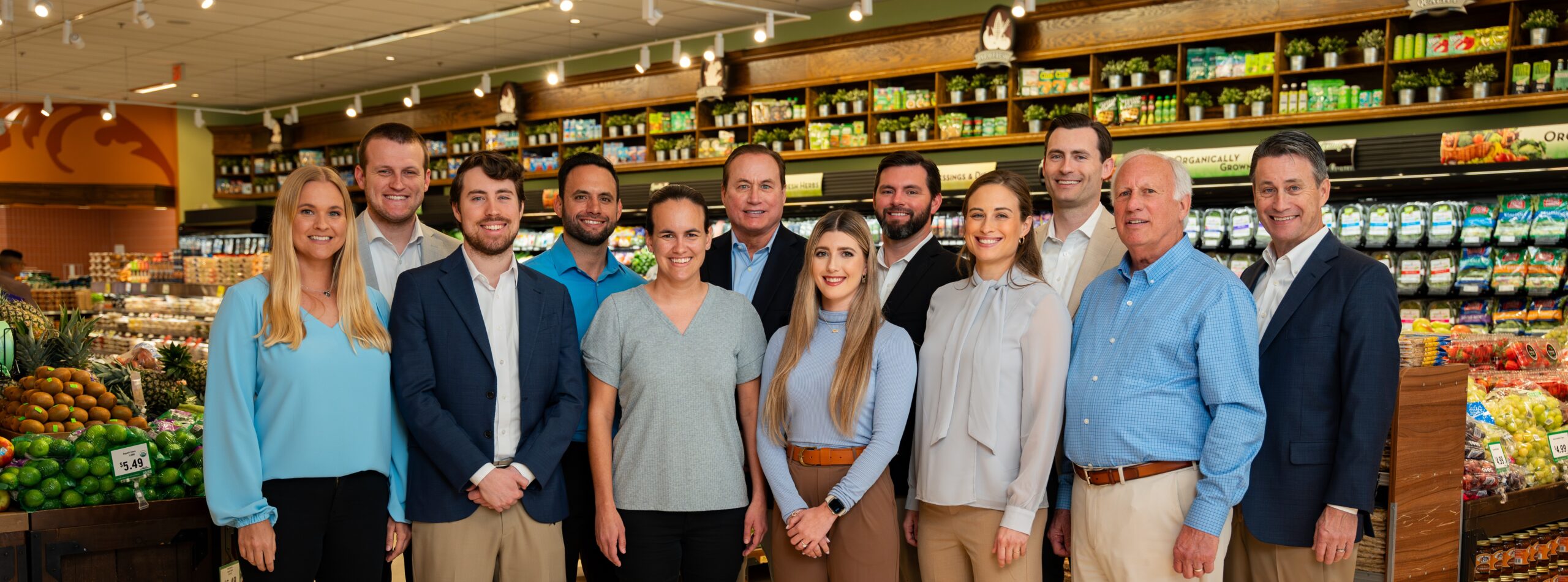 A group of twelve people, dressed in business and casual attire, stand smiling together inside a grocery store with fresh produce and shelves stocked with various food items in the background.