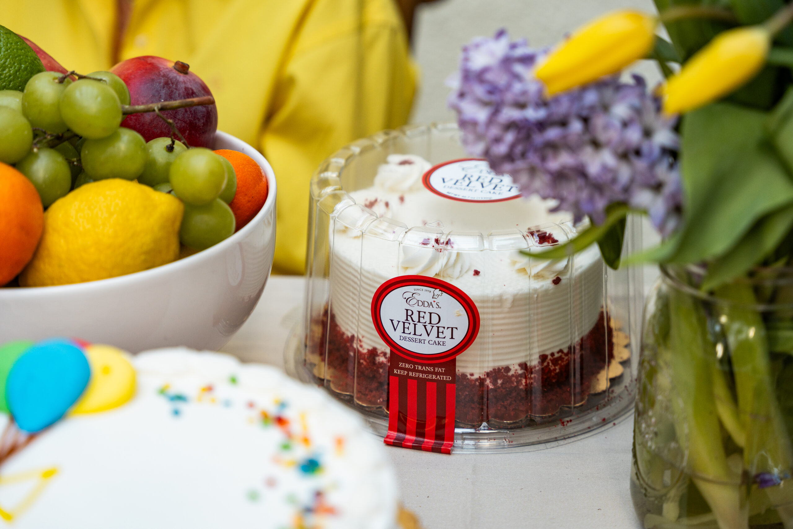 A table with a bowl of assorted fruits, a container of red velvet cake, a partial view of a decorated cake, and a bouquet of yellow tulips and purple flowers.