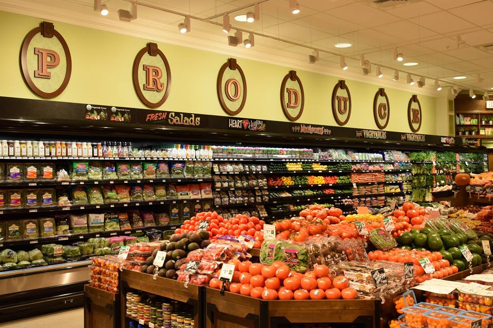 A supermarket produce section with shelves and bins full of fresh fruits and vegetables, including tomatoes, peppers, and avocados. A large “PRODUCE” sign is displayed above the area.