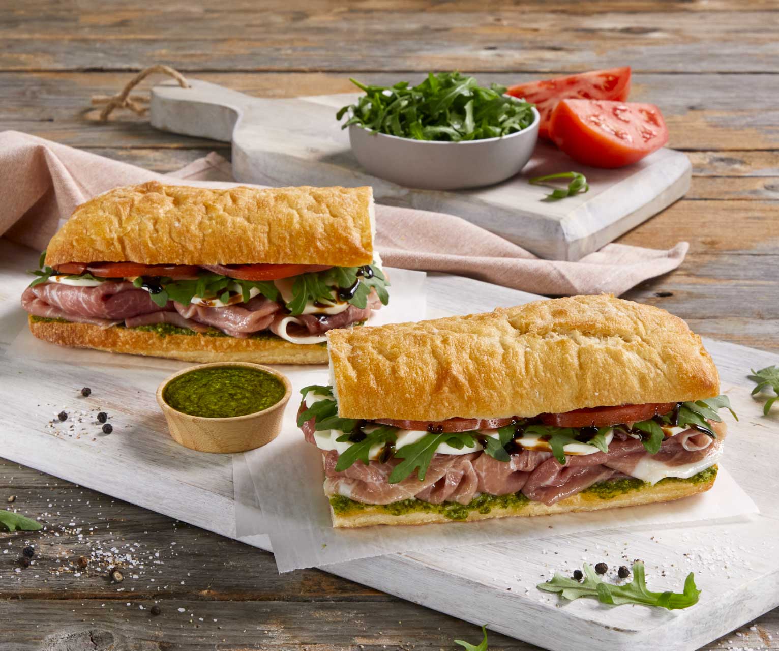 Two ciabatta sandwiches filled with prosciutto, mozzarella, arugula, tomato, and pesto sit on a white board. A bowl of pesto, arugula, and sliced tomatoes are nearby. Order online to enjoy this fresh Italian delight at home!.