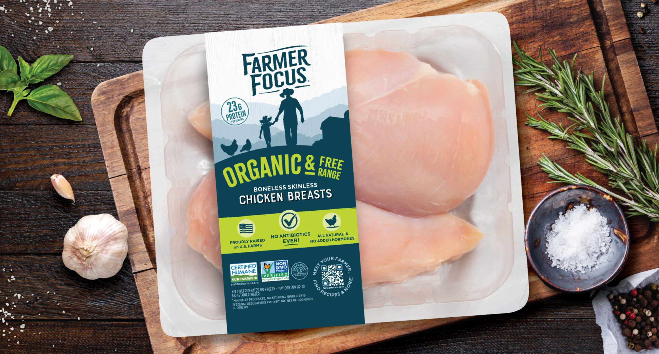 A package of Farmer Focus organic, free-range boneless skinless chicken breasts sits on a wooden table with rosemary, garlic, and peppercorns around it. The label highlights 23g protein, no antibiotics, and USDA organic certification.