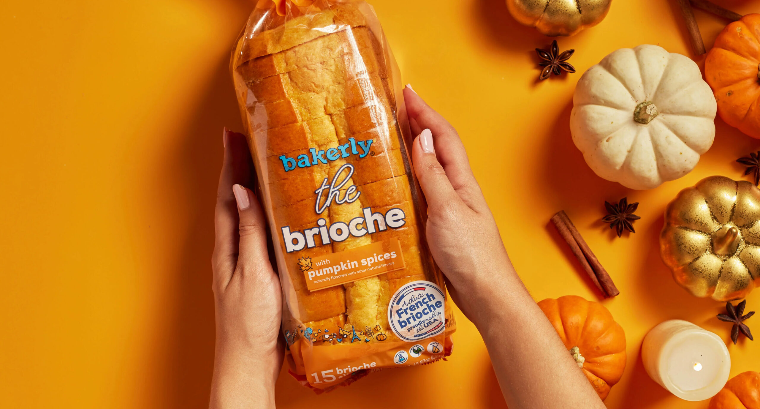 A person holds a loaf of Bakerly pumpkin spices brioche—one of our New Arrivals—on an orange background, surrounded by mini pumpkins, white gourds, cinnamon sticks, and star anise.