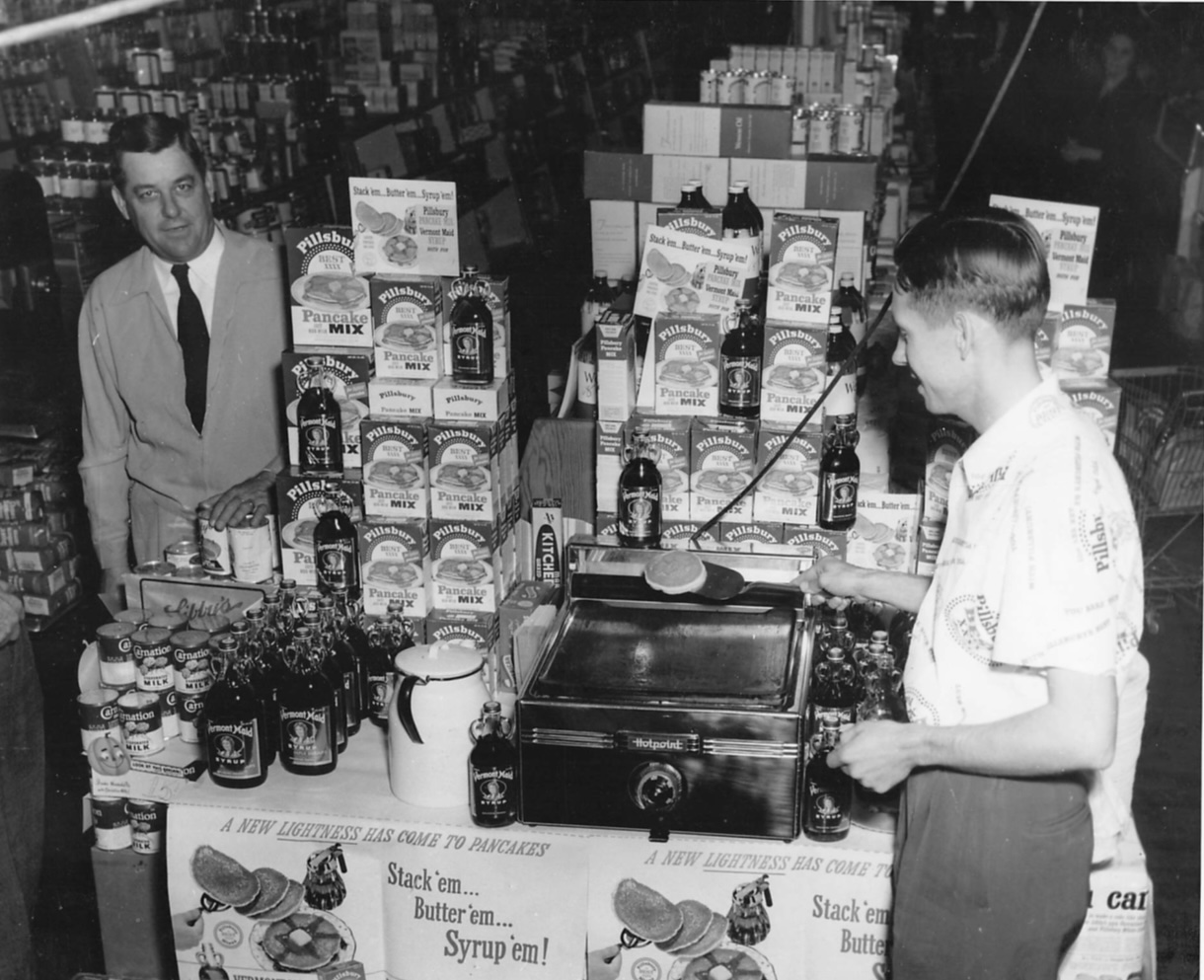 Two men at a supermarket display make pancakes on a griddle, surrounded by boxes of pancake mix and bottles of syrup. One man cooks while the other stands nearby, both dressed in 1950s-style clothing.