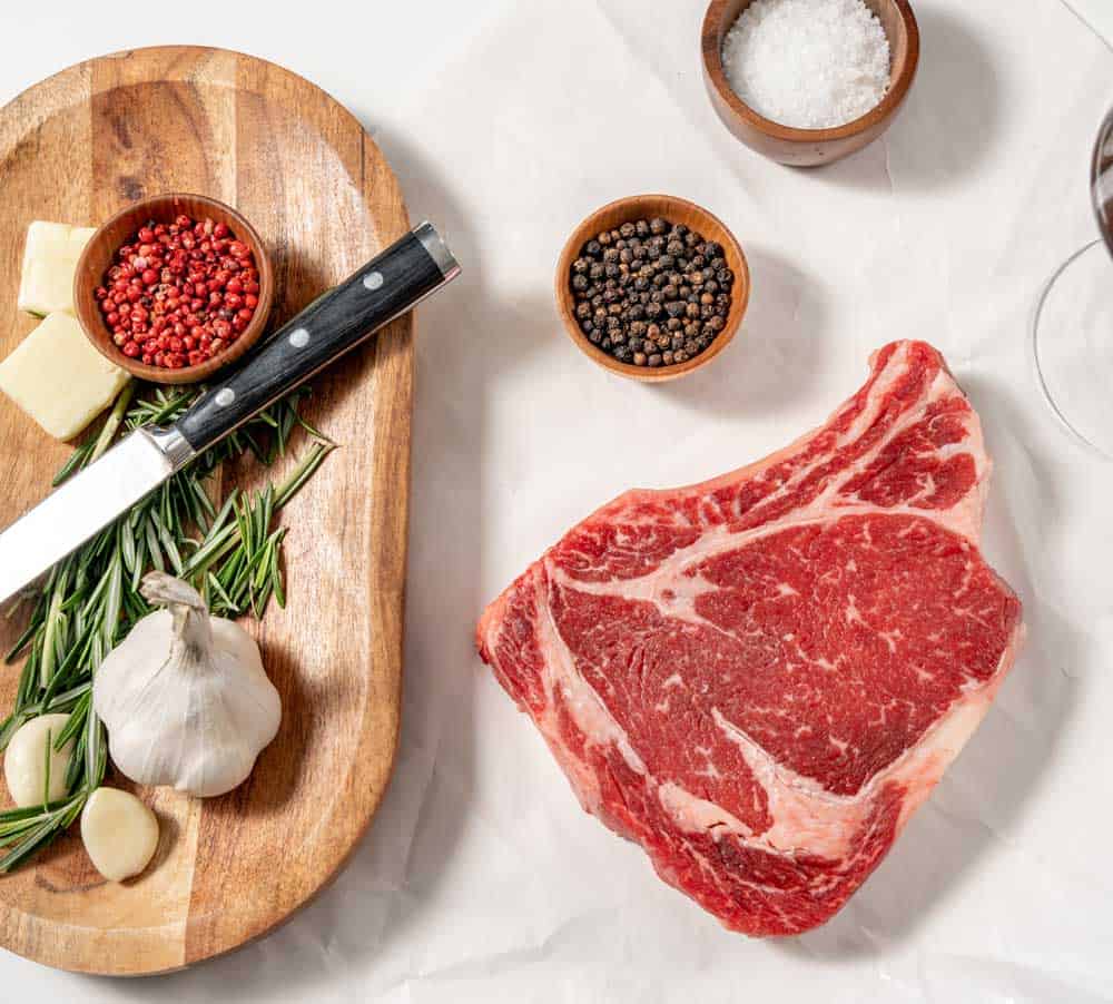 Quality meat ribeye steak on parchment paper next to bowls of black peppercorns and salt. On a wooden board are a knife, garlic, butter, rosemary, and red peppercorns.