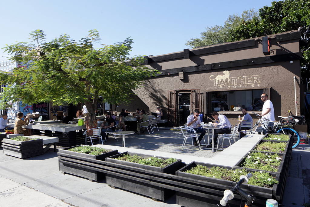 Outdoor seating area of Panther Coffee in Miami with people at tables, surrounded by planter boxes with greenery under a clear sky. The café’s brown building and sign showcase the renowned roasters in the background.