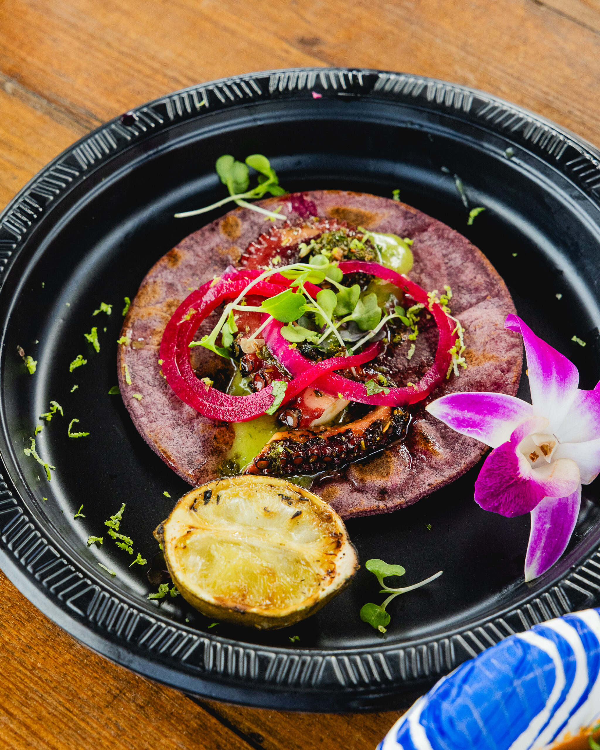 A colorful dish on a black plate features Spanish Octopus Tacos with a purple tortilla topped with greens, onions, and tomato, garnished with microgreens, a grilled lemon half, and a purple orchid, set on a wooden table.
