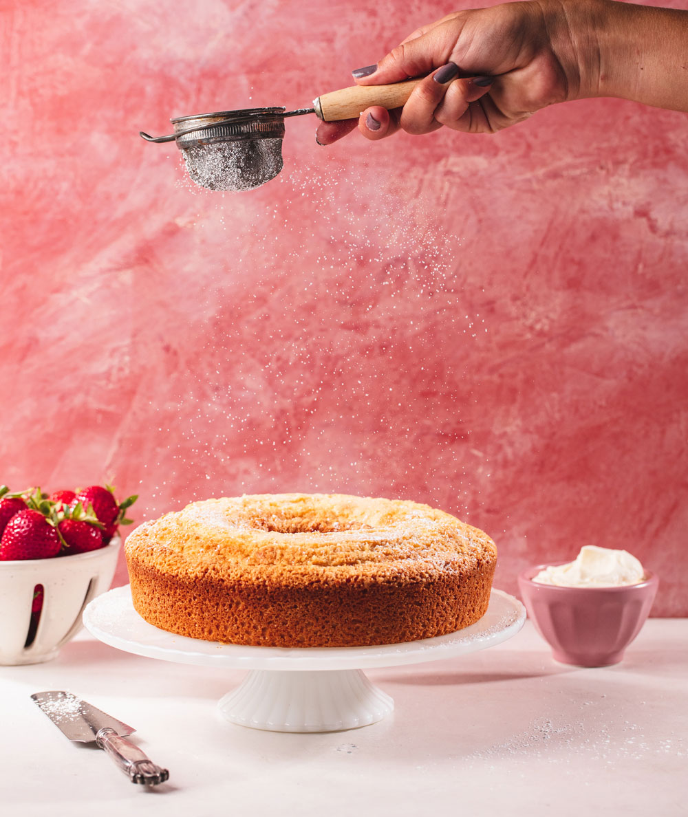 A hand dusts powdered sugar onto a round Grandmother's Pound Cake on a white cake stand. Fresh strawberries, a knife, and a bowl of whipped cream are nearby, all set against a pink textured background.