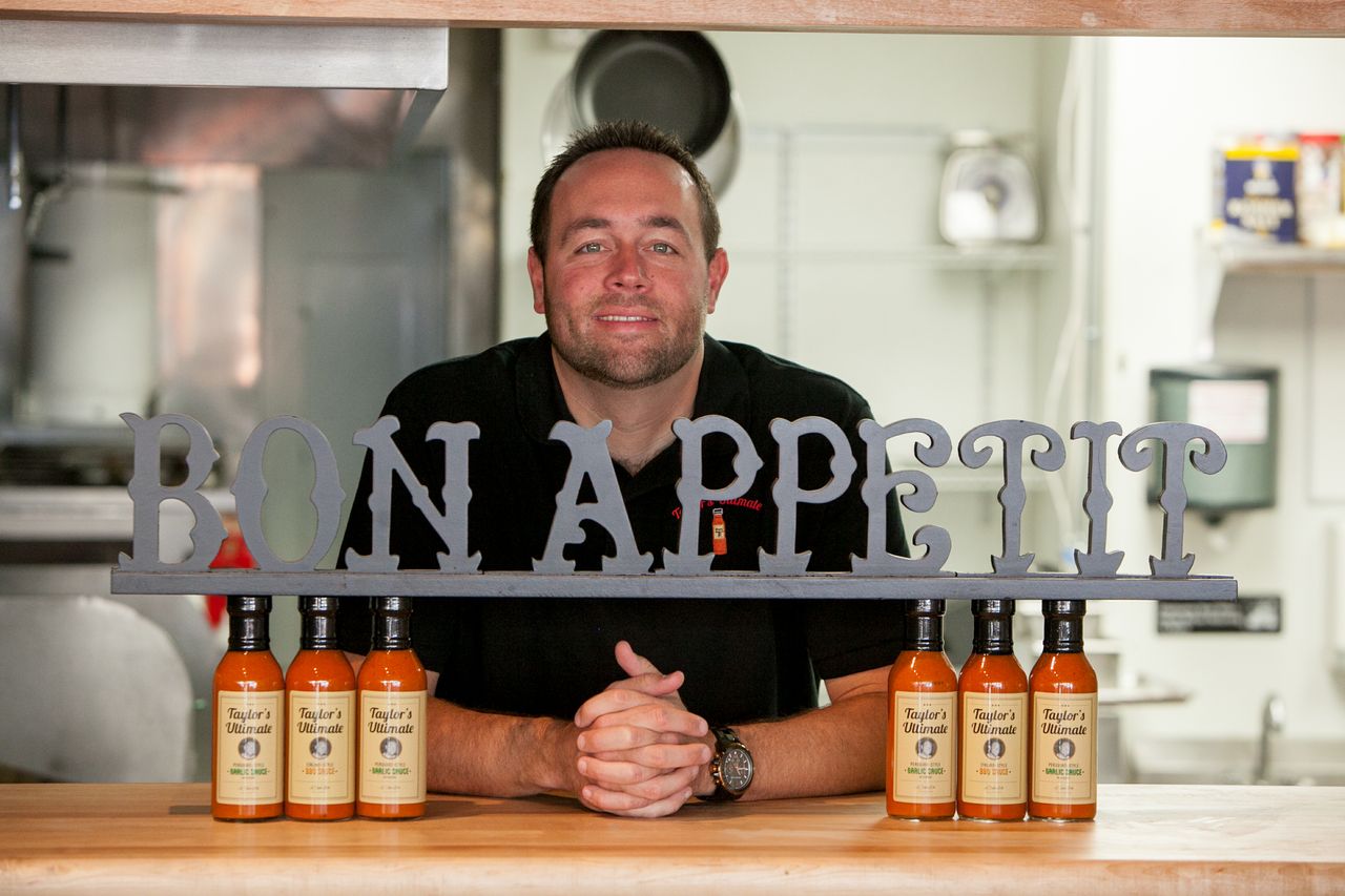 A man in a black shirt smiles behind a counter with a decorative BON APPETIT sign and six bottles of Taylor’s Ultimate Sauces arranged in front of him, in a kitchen setting.