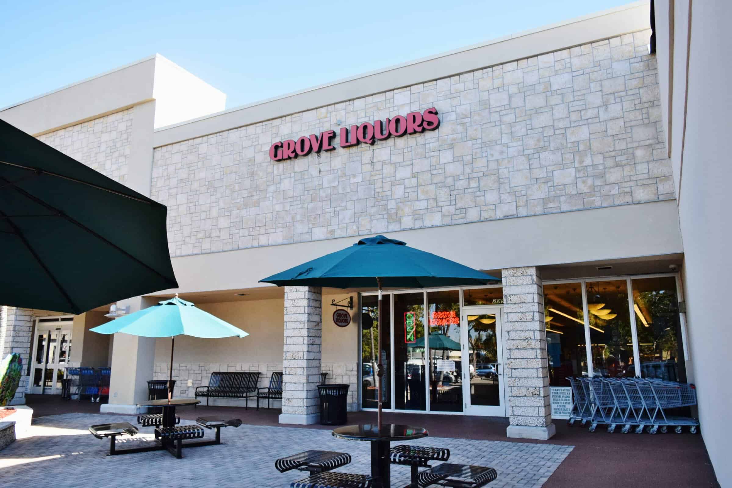 Outdoor seating area with green umbrellas and picnic tables in front of a store called Grove Liquors. The building has a stone facade and large glass windows. Shopping carts are visible near the entrance.