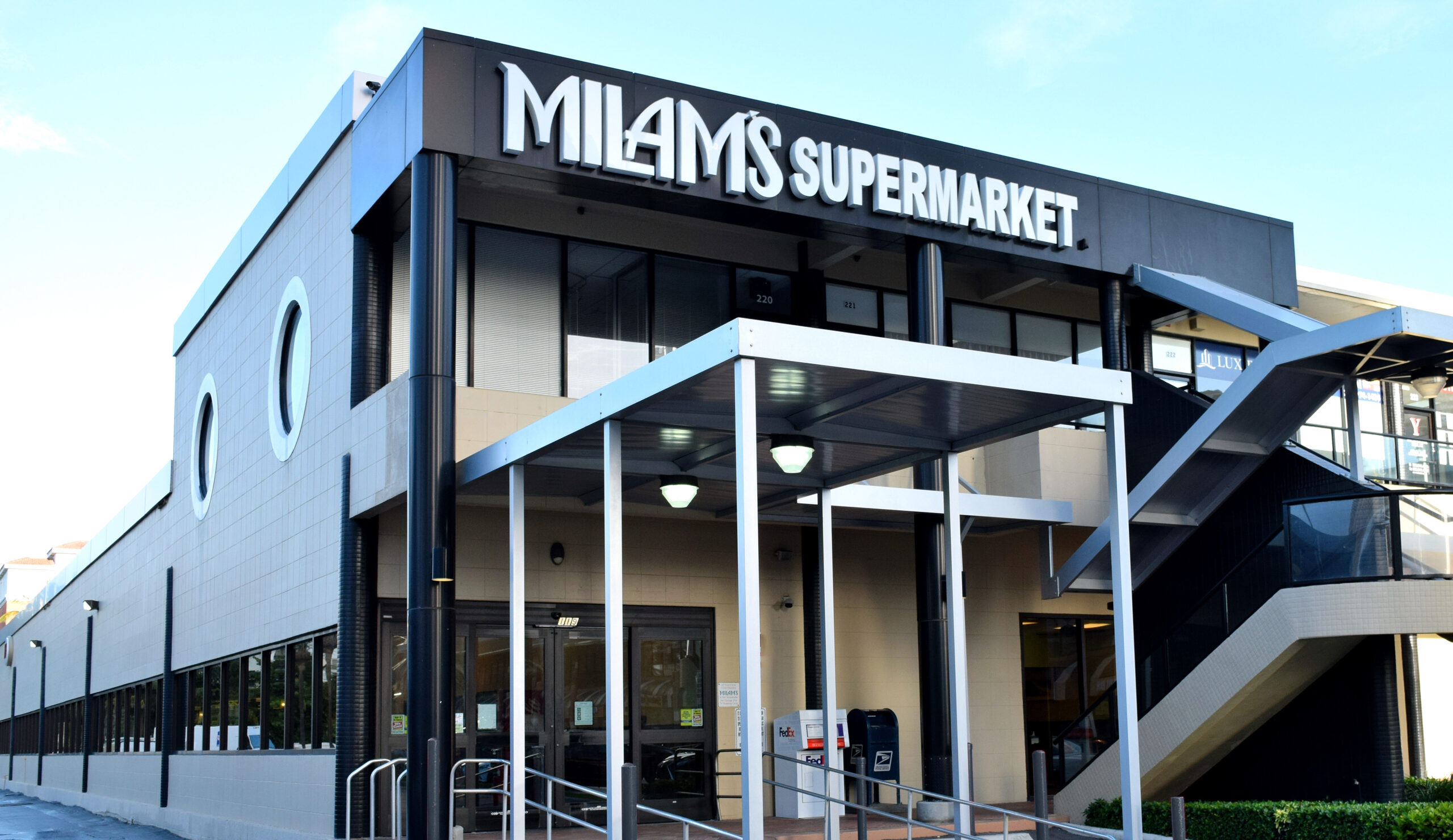 The image shows the front entrance of Milam’s Supermarket, a two-story building with large windows and a black and white sign above the entrance. A covered walkway leads to glass double doors.