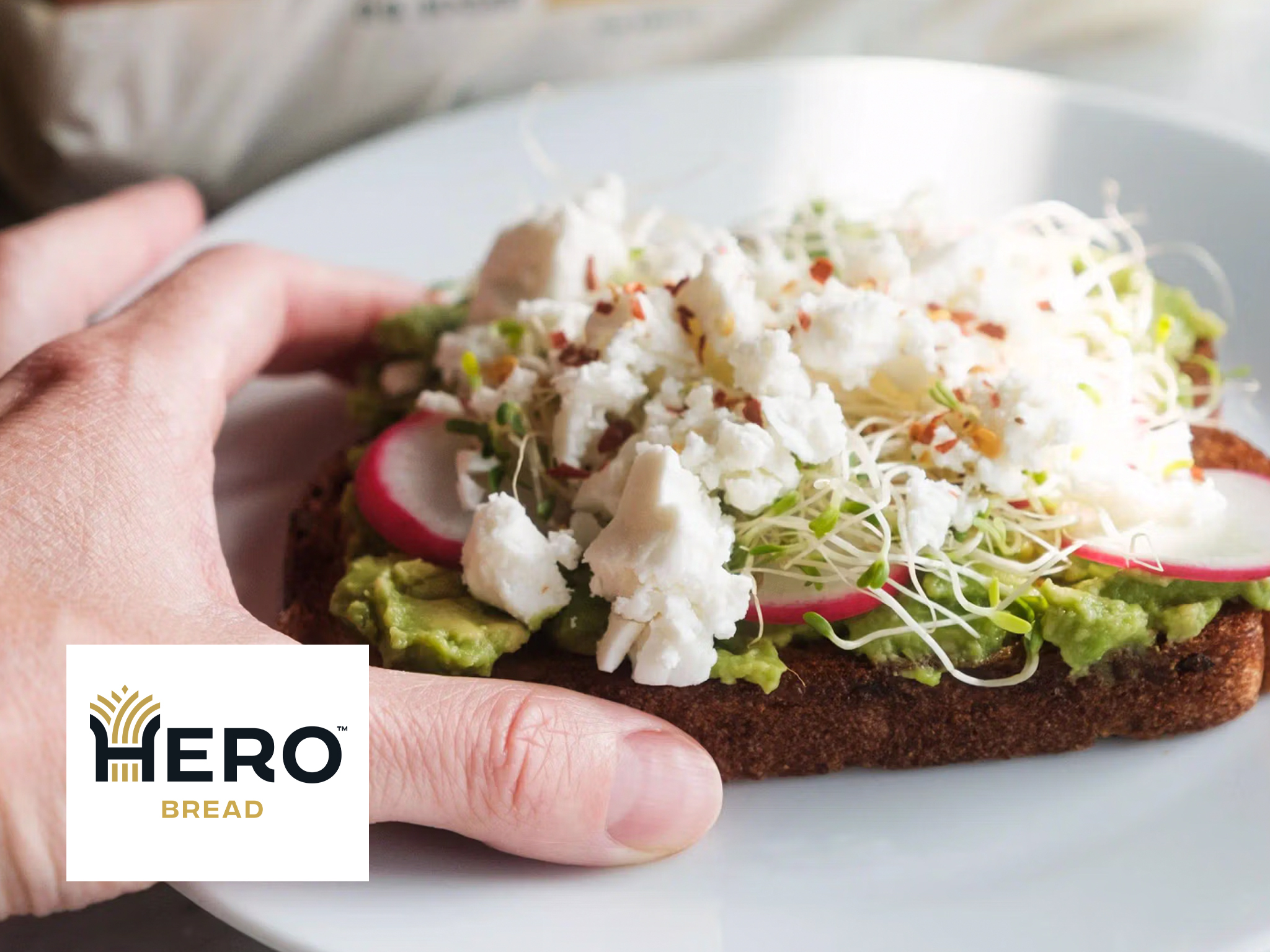 A hand holds a slice of Avocado Toast topped with radish, sprouts, and crumbled cheese on a white plate. The Hero Bread logo is visible in the lower left corner.