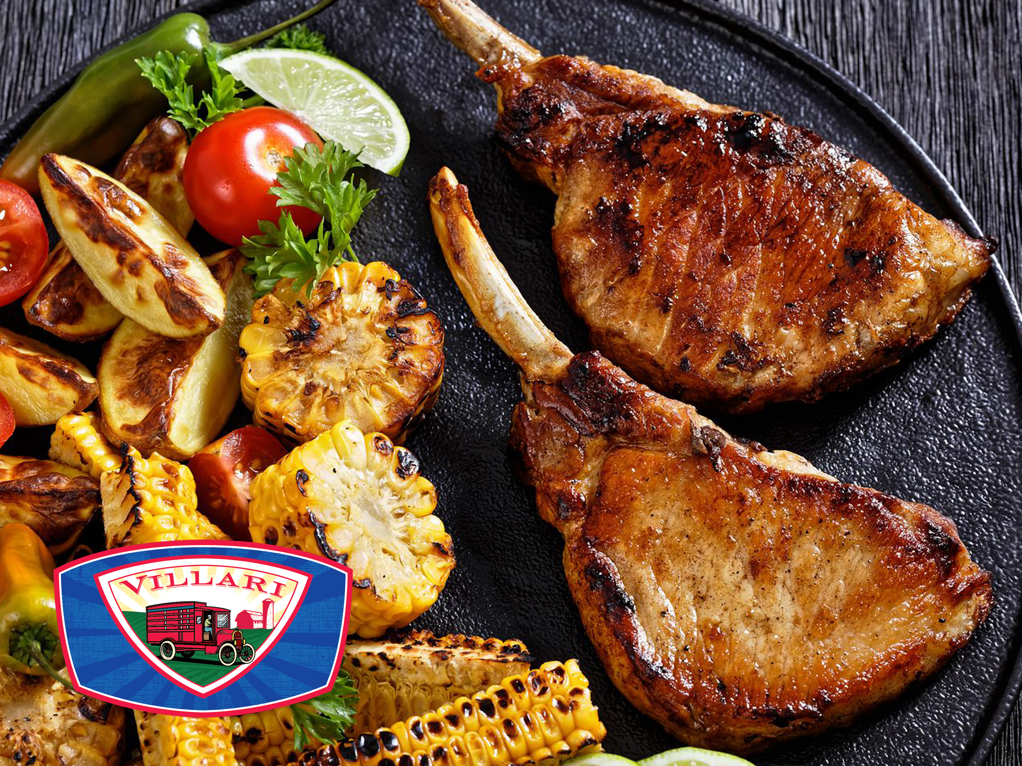 Two grilled pork chops, including a savory Tomahawk Chop, are served on a black plate with corn, roasted potatoes, cherry tomatoes, lime slices, and a green chili pepper. The Villari logo is in the bottom left corner.