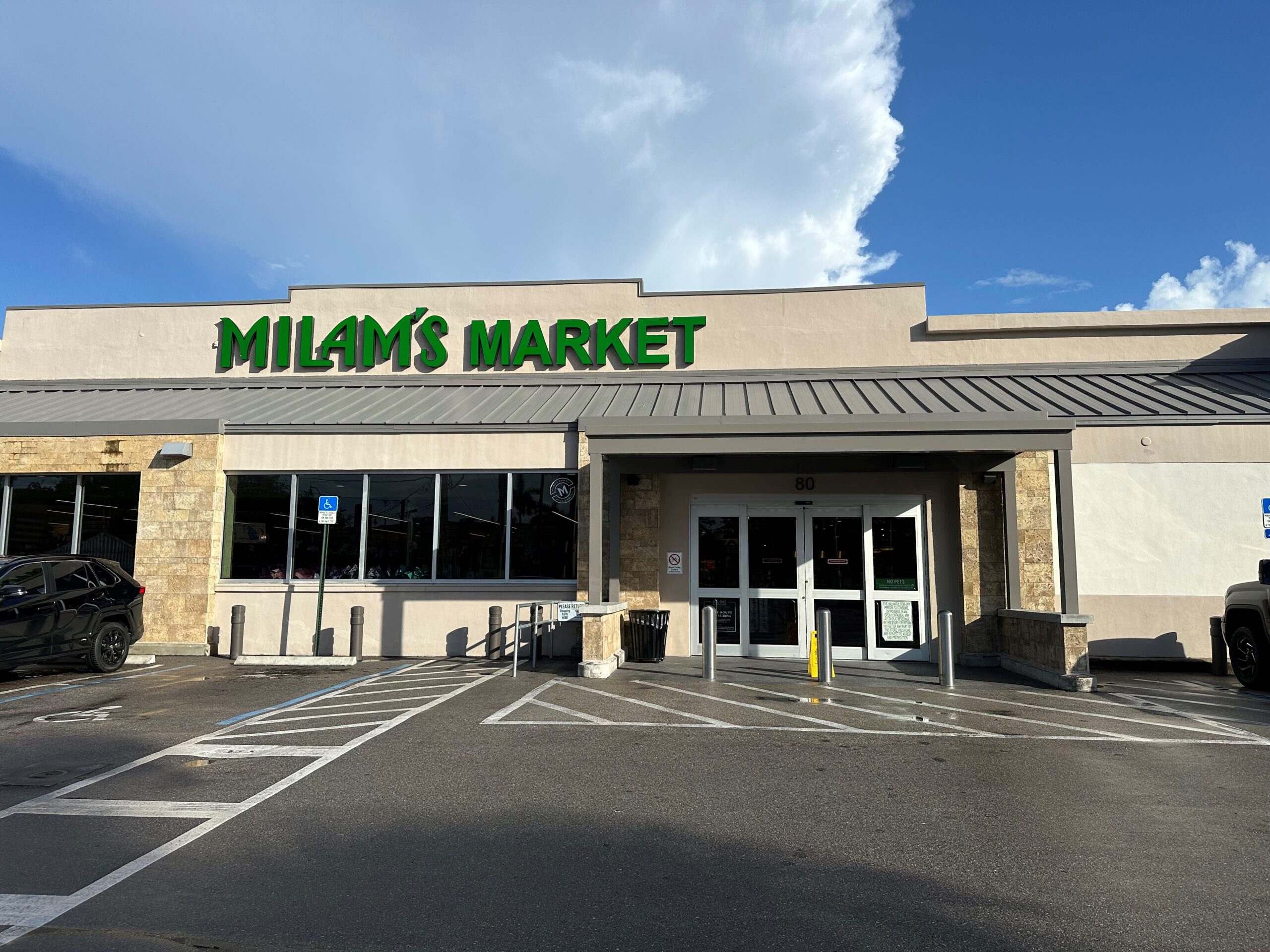 Milam’s Market storefront in Miami Springs features green signage, large front windows, automatic glass doors, and accessible parking spaces under a bright blue sky with clouds.