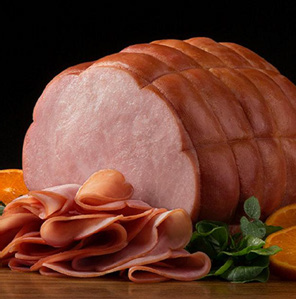 A large, sliced ham sits on a wooden surface, surrounded by thin slices of ham, green leafy garnish, and orange slices, all set against a black background.