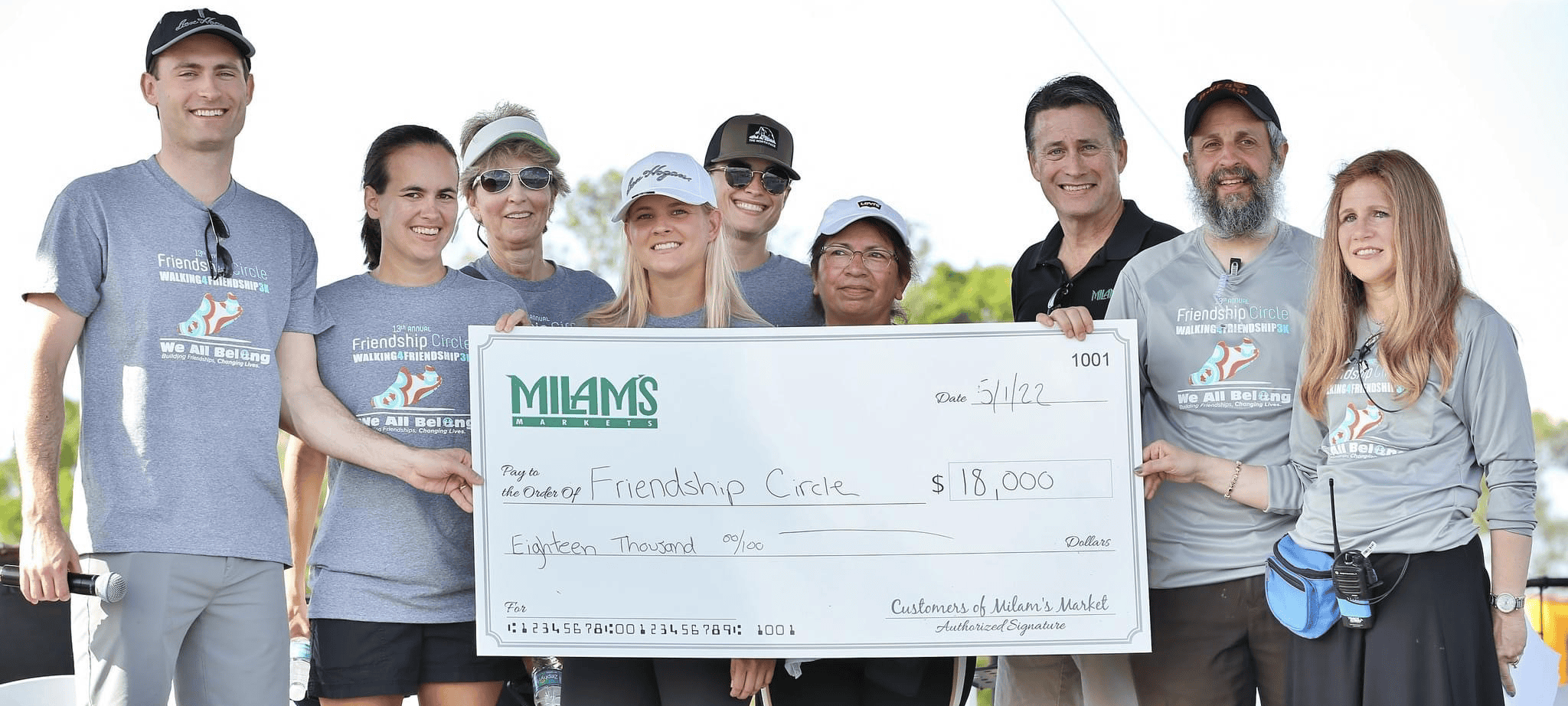 A group of smiling people in athletic wear hold a large check for $18,000 made out to Friendship Circle, standing outdoors on a sunny day—celebrating generous community donations.