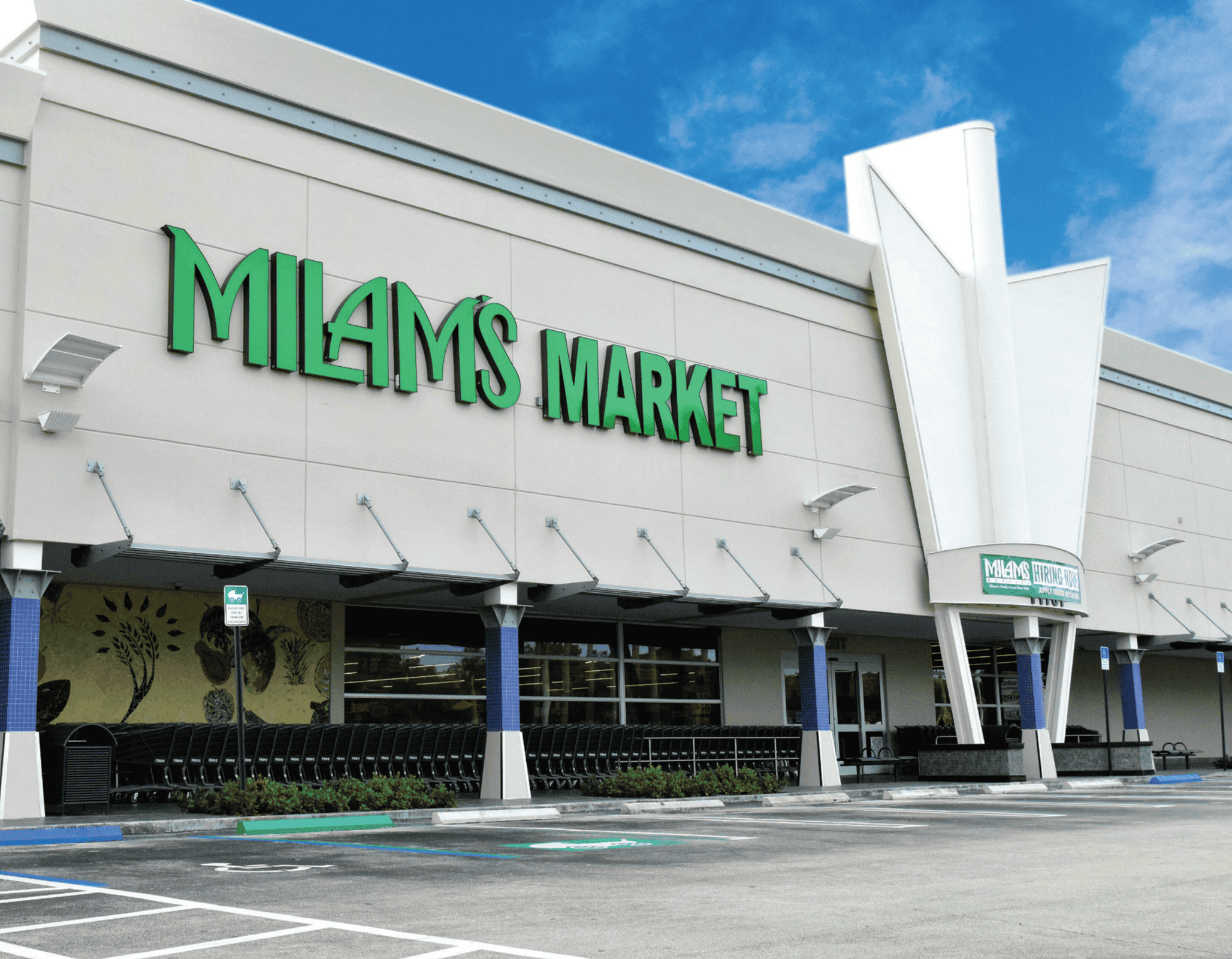 The exterior of Milams Market, a grocery store with a modern white facade and green signage, set against a blue sky with some clouds; shopping carts and empty parking spaces are visible in front.