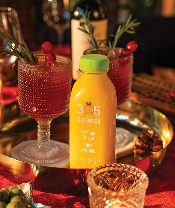 A bottle of 305 Squeezed Orange Ginger juice sits on a gold tray, surrounded by festive glassware filled with sparkling mocktails garnished with rosemary and cranberries, on a vibrant red tablecloth.