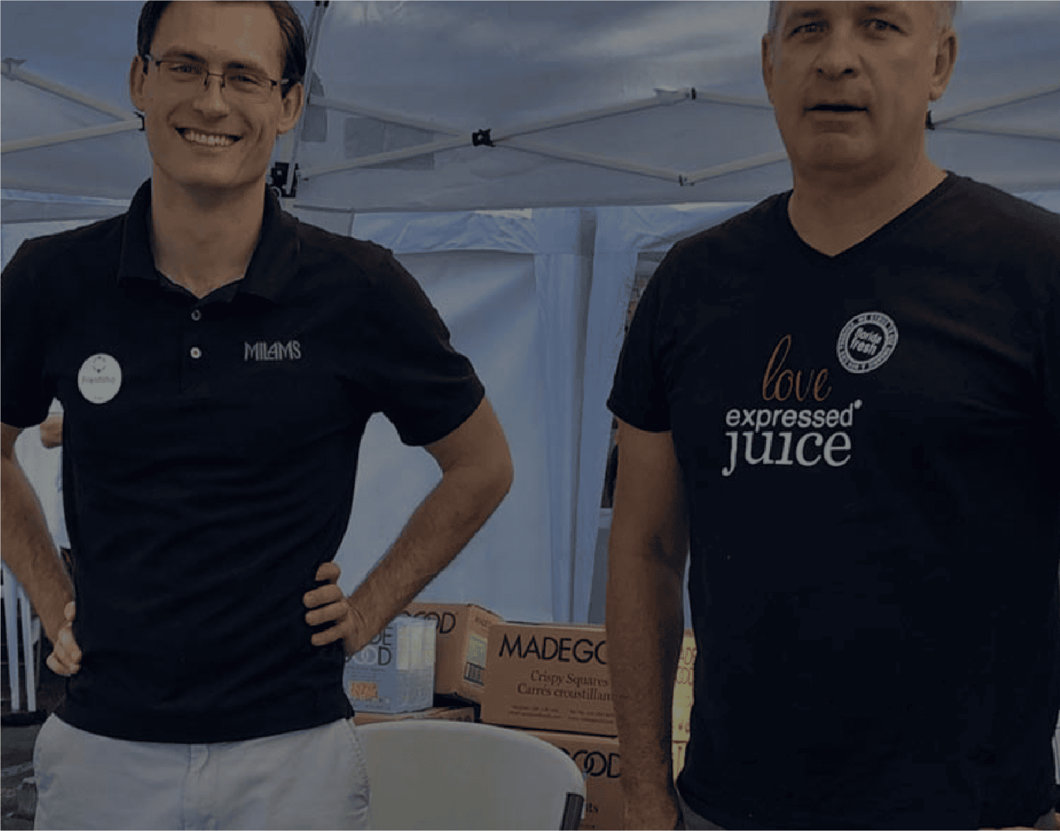 Two men stand side by side under a canopy. One wears glasses and a black polo with a name tag, smiling. The other wears a black t-shirt with "love expressed JUICE." Behind them, boxes for community donations are stacked.