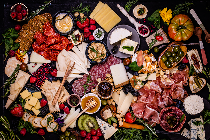 A colorful charcuterie board topped with assorted cheeses, cured meats, crackers, fruits, nuts, dips, herbs, and edible flowers, arranged artfully on a dark surface.