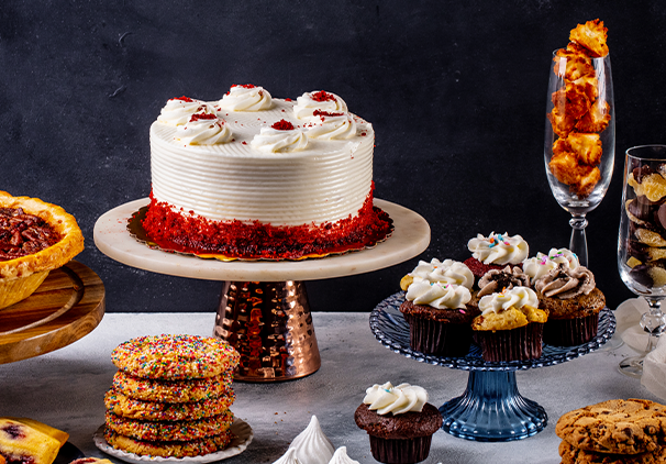 A dessert table featuring a red velvet cake with white frosting, assorted cupcakes, cookies, meringues, a pecan pie, and a champagne glass filled with coconut macaroons against a dark background.