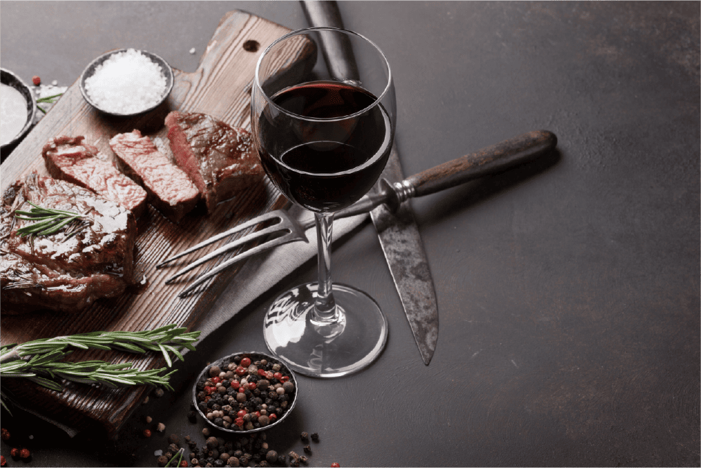A glass of red wine sits beside a cutting board with sliced quality meat steak, rosemary sprigs, a fork, a knife, coarse salt, and a small bowl of mixed peppercorns on a dark surface.