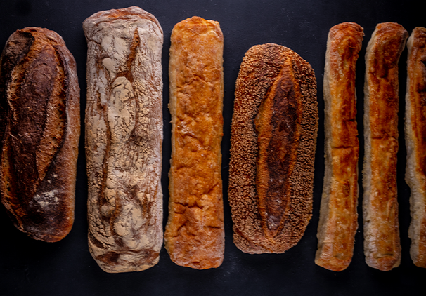 Seven artisanal bread loaves of various shapes and crust colors are arranged vertically on a dark background, showcasing different textures and baking styles.