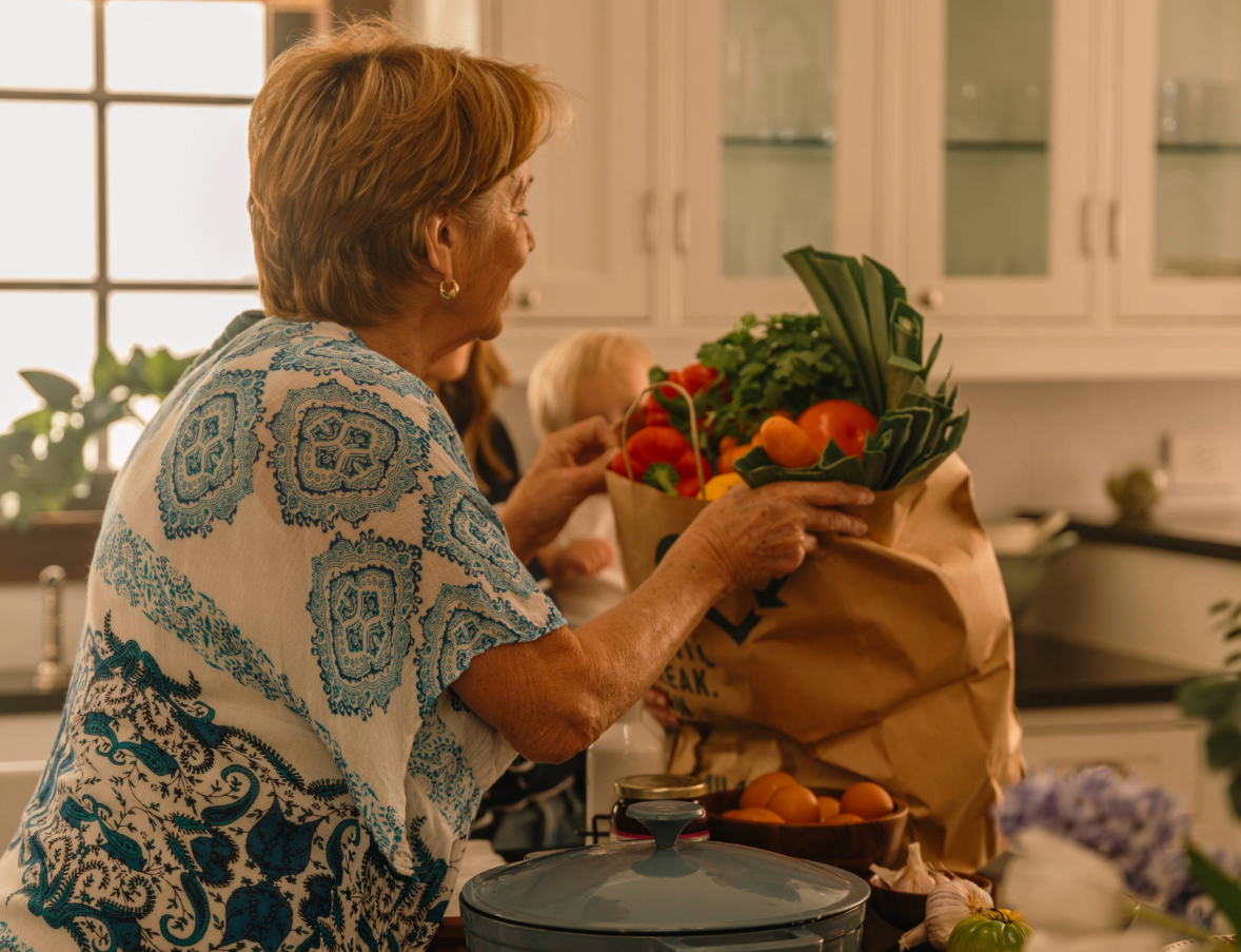 An older woman in a kitchen holds a paper grocery bag filled with fresh vegetables, including tomatoes, leeks, and parsley. Sunlight streams in through a window, and kitchen items are visible on the counter.